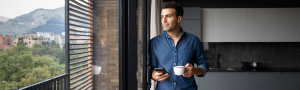 Eine Person, steht in einer modernen Küche und hält eine Tasse Kaffee in der einen Hand und ein Smartphone in der anderen. Im Hintergrund sind große Fenster mit Blick auf eine Landschaft mit Bäumen und Bergen zu sehen.