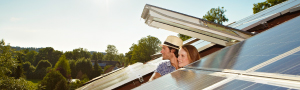Zwei Personen, schauen aus einem Dachfenster heraus auf ein Dach mit Solarpaneelen. Im Hintergrund sind Bäume und ein klarer Himmel zu sehen.