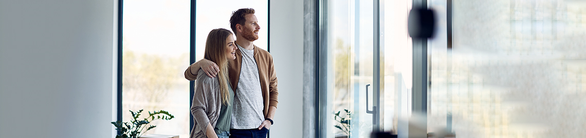 Junges umarmtes Paar, das durch das Fenster ihrer neuen Wohnung schaut.